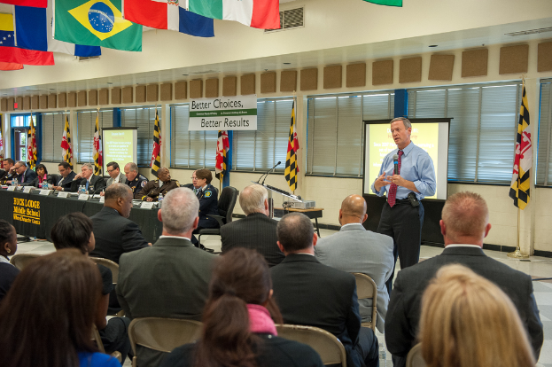 Eine Gruppe von Menschen, die auf Stühlen sitzen und einem Redner mit Mikrofon zugewandt sind, mit einem Projektionsschirm und einem Tisch mit einem Projektor dahinter, einer Reihe von Menschen an einem Tisch mit Wasserflaschen, Fahnenmasten, hängenden Fahnen, abgedeckten Fenstern und einer Tür auf der linken Seite.
