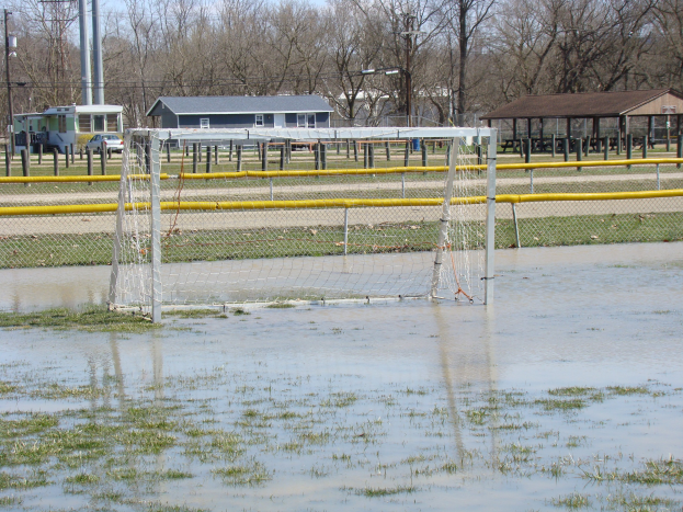 Ein Fußballtor steht in der Mitte eines überfluteten Feldes mit Gras und Wasser, mit Hütten, Pfählen, Bäumen, Fahrzeugen und einem klaren blauen Himmel im Hintergrund.