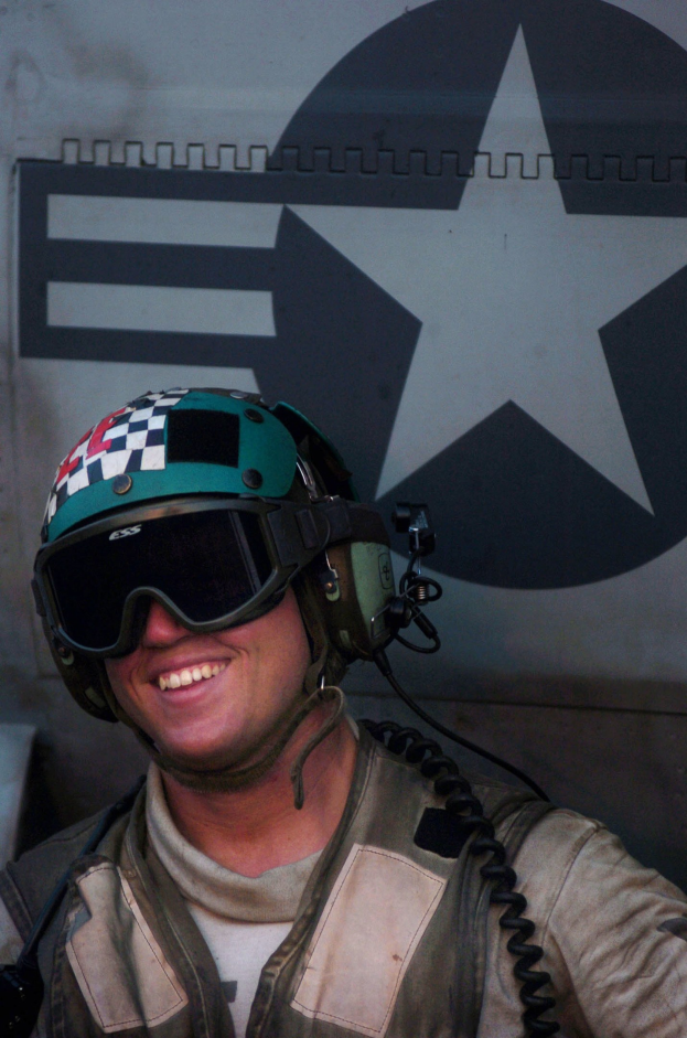 A smiling pilot in uniform and helmet sits in front of a plane, with a logo visible on the wall behind him.