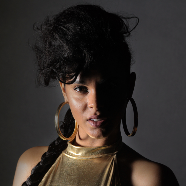 Eine Frau in einem goldenen Kleid und großen Ohrringen steht selbstbewusst vor einer Wand, ihr glatter Bob-Haarschnitt und das natürliche Make-up unterstreichen ihren entschlossenen Gesichtsausdruck, während sie direkt in die Kamera schaut.