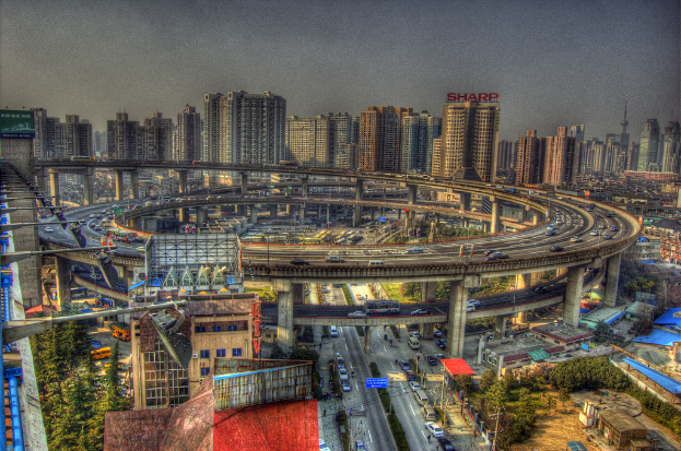 Eine Stadtansicht mit einem zentralen Ringverkehr mit Fahrzeugen, einer unteren Straße mit Autos und Fahrrädern, zahlreichen Hochhäusern im Hintergrund, einem Himmel oben, Bäumen auf der linken unteren Seite und kleinen Häusern, die über die Stadt verteilt sind.