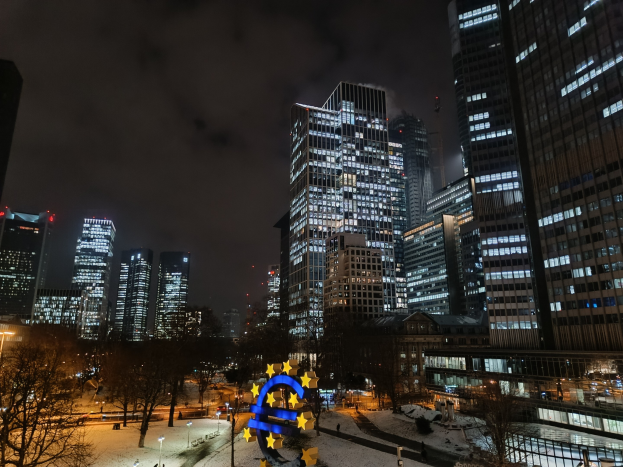 Europäische Zentralbank-Hauptquartier in Frankfurt, Deutschland bei Nacht mit beleuchteten Gebäuden, Bäumen, Straßenlaternen, Lichtern und Fußgängern unter einem bewölkten Himmel.
