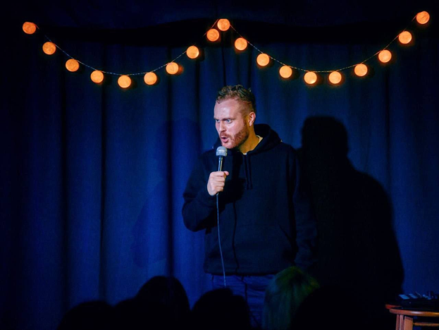 A man stands on a brightly lit stage holding a microphone, with a table of objects to his right and an audience visible below.