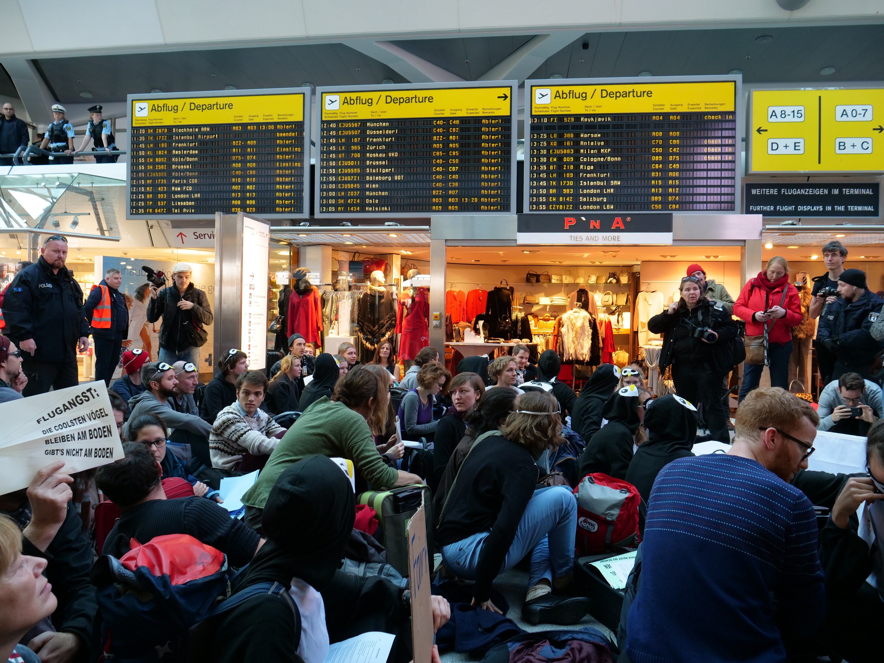 Eine große Gruppe von Menschen in einem Flughafen, einige sitzen mit Taschen und Papieren, andere stehen, mit Texttafeln und Schaufensterpuppen im Hintergrund, was auf eine Demonstration hinweist.