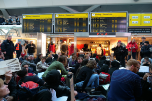 Eine große Gruppe von Menschen in einem Flughafen, einige sitzen mit Taschen und Papieren, andere stehen, mit Texttafeln und Schaufensterpuppen im Hintergrund, was auf eine Demonstration hinweist.
