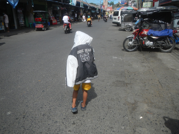 Ein junger Junge geht eine Straße voller Motorräder und anderer Fahrzeuge entlang, flankiert von Gebäuden und Bäumen unter einem klaren blauen Himmel.
