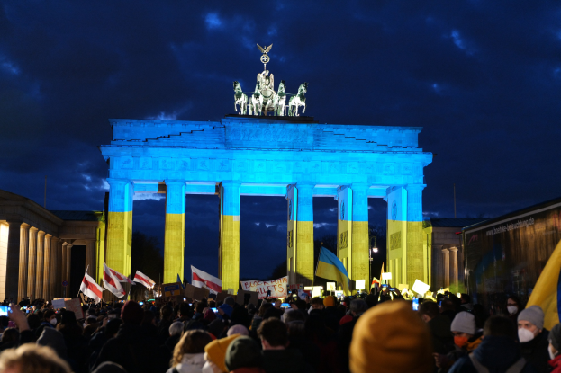 A crowd of people stands in front of the Brandenburg Gate in Berlin, holding flags and placards, with a banner on the right side displaying protest-related text.