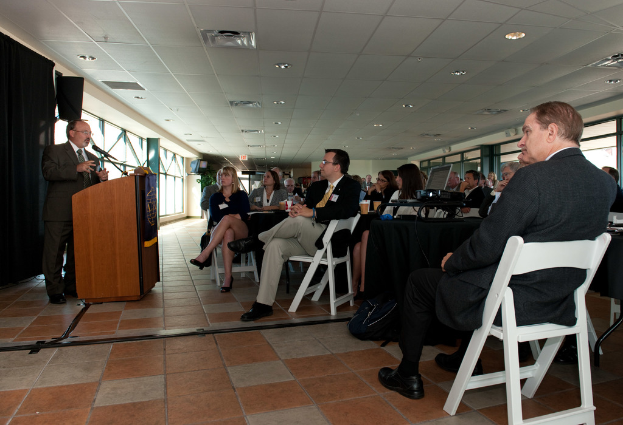 Eine Gruppe von Menschen, darunter Männer und Frauen, sitzt einem Mann gegenüber, der an einem Podium steht und in ein Mikrofon spricht, mit Fenstern im Hintergrund.