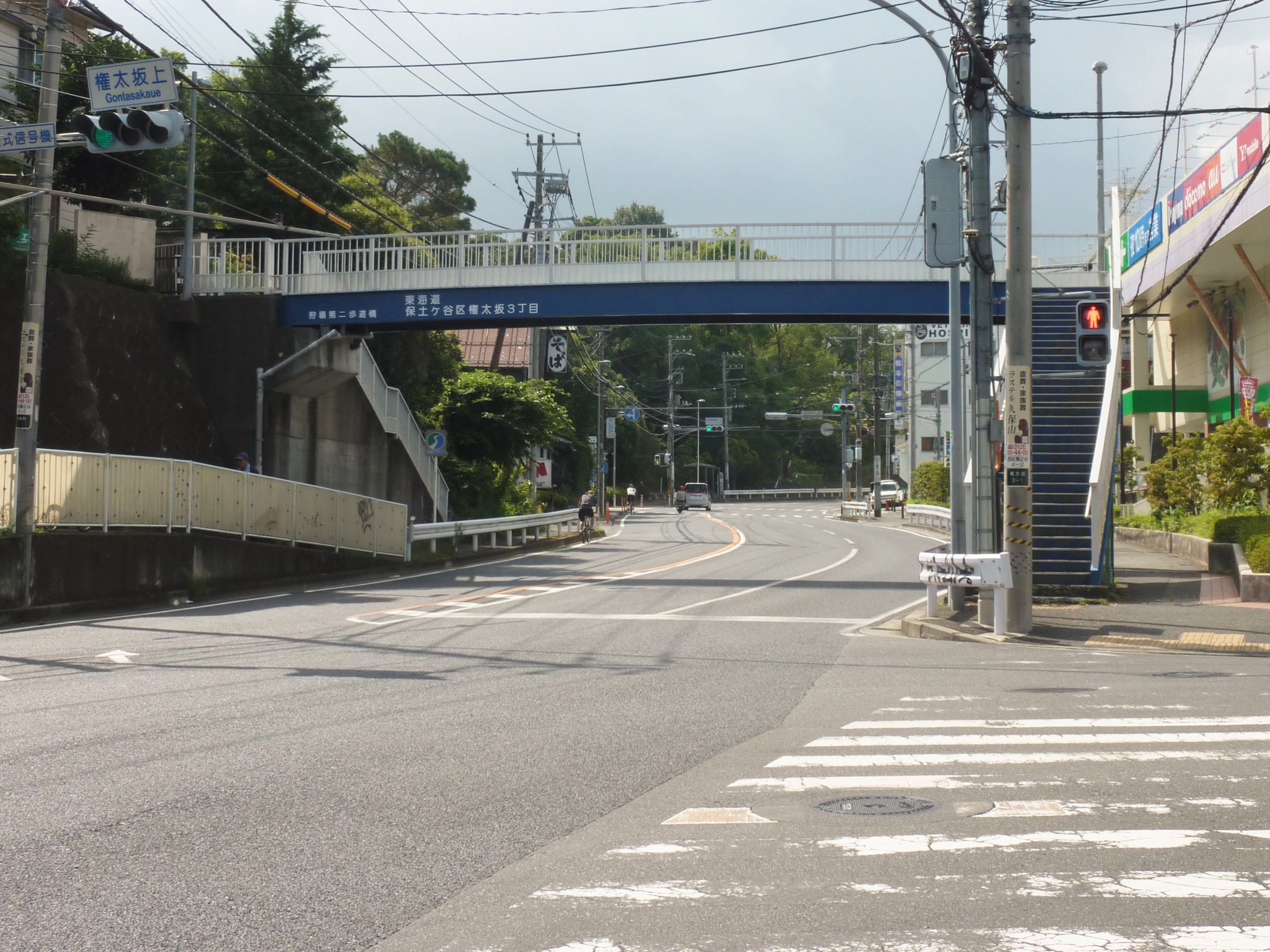 Eine Stadtstraße mit einer Fußgängerbrücke darüber, Fahrzeuge auf der Straße, Strommasten mit Drähten, Verkehrsampeln, Schilder, Gebäude mit Fenstern, Bäume und ein Himmel im Hintergrund.