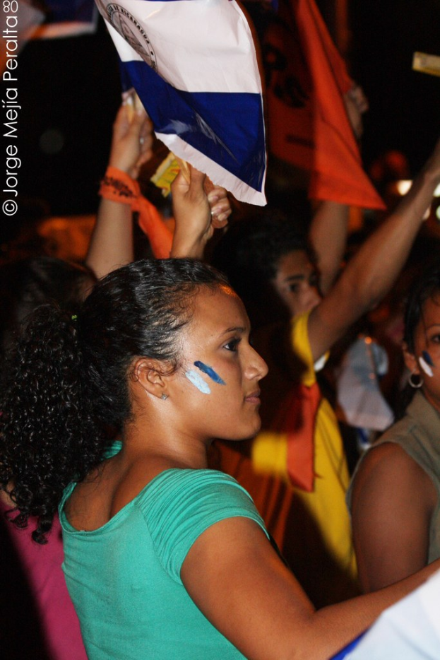 Frauen in grünen T-Shirts halten eine weiße Abdeckung während einer Protestveranstaltung, mit mehreren Mädchen, die dahinter stehen.