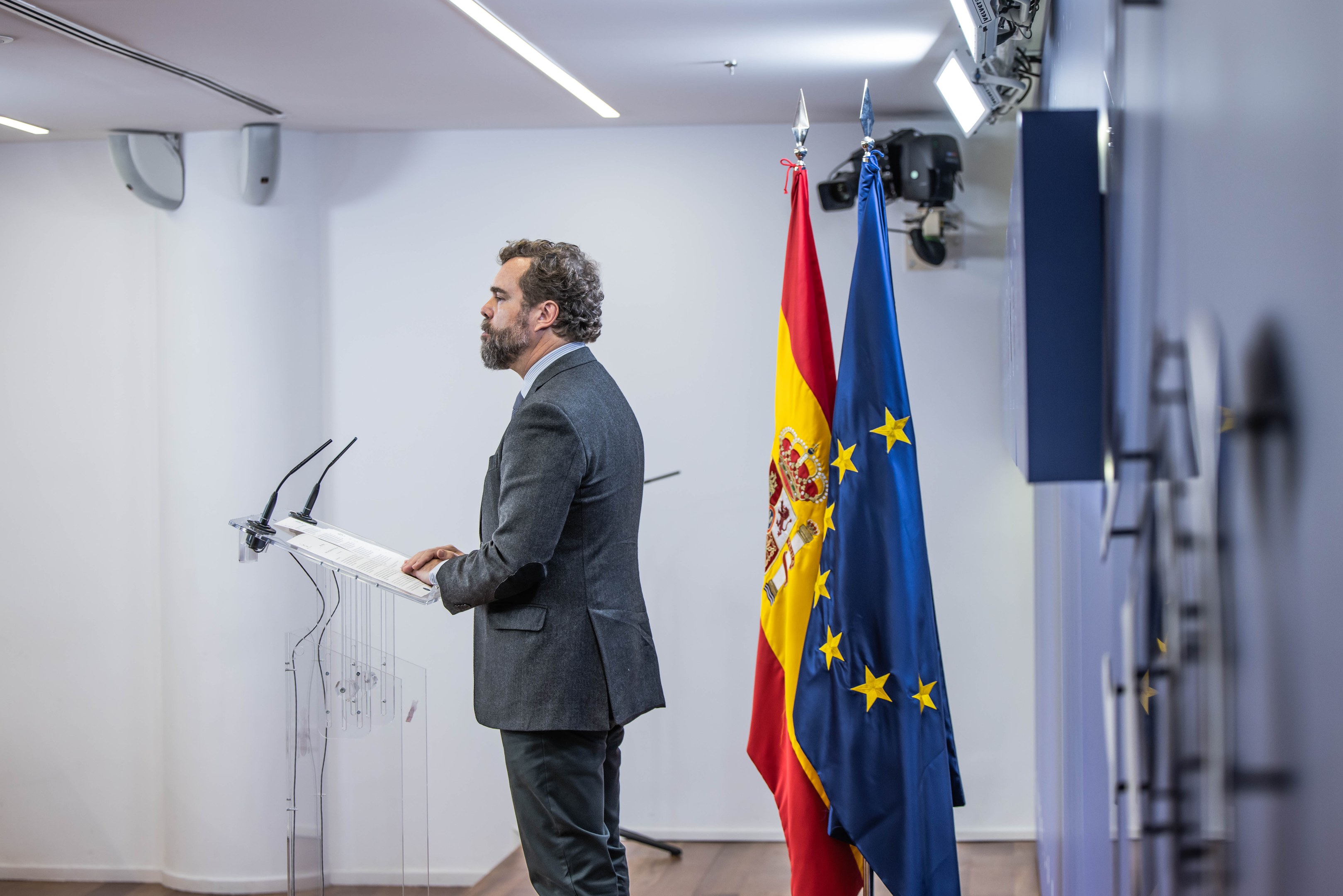 A man stands at a podium holding a paper, speaking into microphones, with two flags behind him and a door to the right, in a room with wall-mounted lights and objects.