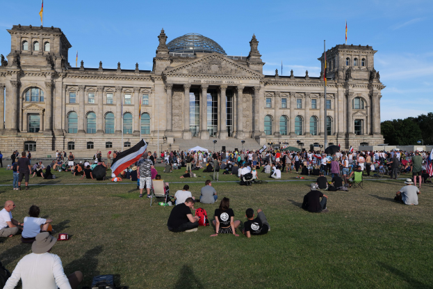 Eine große Gruppe von Menschen sitzt und steht auf einem grünen Feld vor dem Reichstaggebäude in Berlin, Deutschland, mit Taschen und Gegenständen, die herumliegen, Bäumen und einem klaren blauen Himmel im Hintergrund.