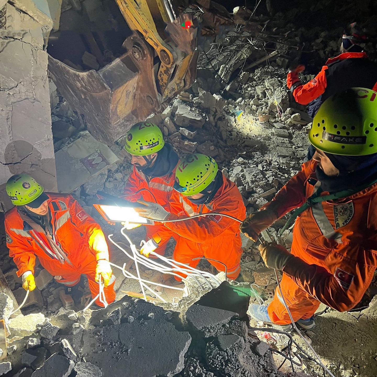 Gruppe von Menschen in orangen Uniformen und grünen Helmen bei der Arbeit an Schutt von einem eingestürzten Gebäude mit einem Kran im Hintergrund.