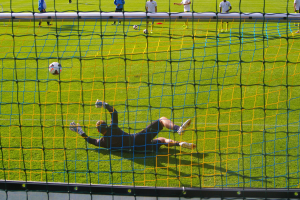 Ein Fussballer wehrt einen Ball vor einem Tor ab, mit einem Netz im Vordergrund und einigen Menschen im Hintergrund, sowie einigen Bällen auf dem Boden verteilt.