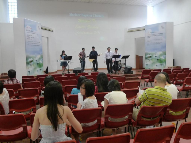 Eine Bühne mit zwei Gitarristen in der Mitte und anderen Musikern in den Ecken, die singen und Instrumente spielen, mit Bannern an den Seiten und einem Publikum davor, das Papiere und Bücher hält.