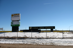 Eine Straße mit Schnee, ein Zaun mit Text, eine Wand, Laternenpfähle, Bäume und ein klarer Himmel, mit einem Schild, das "Chinle High School" im Vordergrund anzeigt.