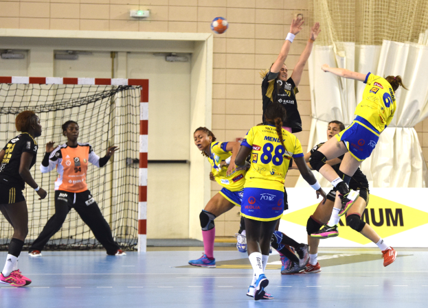 Eine Gruppe von Frauen beim Handballspielen auf einem Platz, mit einem Ball in der Luft, einem Tor mit Netz, einer Fahne mit Text, einem Vorhang und einer Wand im Hintergrund.