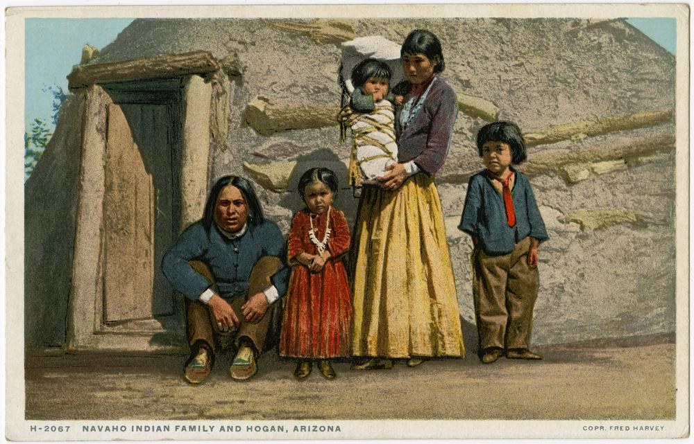 Black-and-white postcard of a Navajo family standing in front of a wooden house with a door, trees, and a clear sky, labeled "Navajo Indian Family and Hogan, Arizona" at the bottom.
