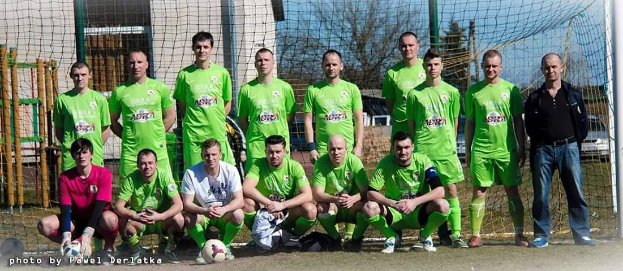 Eine Gruppe von Männern in grünen T-Shirts steht auf einem Fußballfeld mit einem Ball davor, einige hocken sich hin, in der Nähe eines Torpfostens mit Bäumen, Gebäuden und Fahrzeugen im Hintergrund.