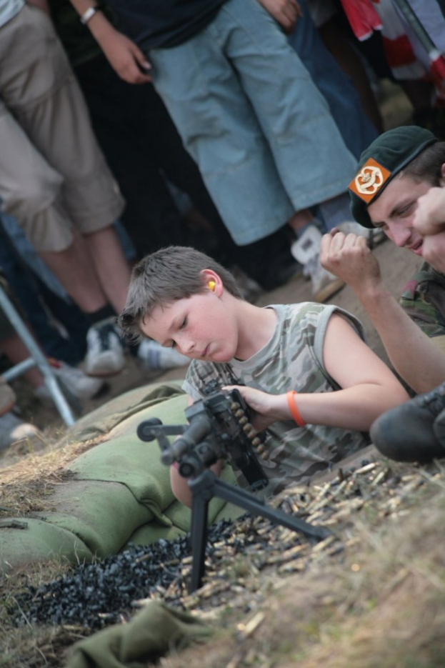 Ein junger Junge sitzt auf dem Boden und hält eine Maschinenpistole, umgeben von einer Gruppe von Menschen im Hintergrund.