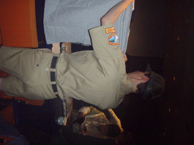 Ein Polizeibeamter hält ein T-Shirt in den Vordergrund, mit einem Jungen im Hintergrund, der ein Buch und einen Becher hält und in der Nähe eines Schreibtisches und Lampen steht.