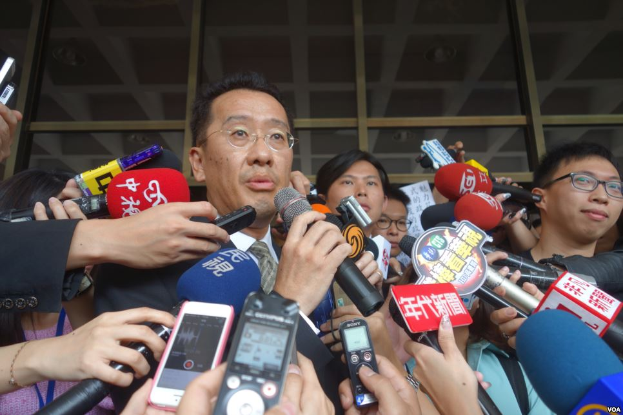 Ein Mann im Anzug und Krawatte, der eine Brille trägt, steht umgeben von Reportern mit Mikrofonen und Handys, vor einer Glaswand.