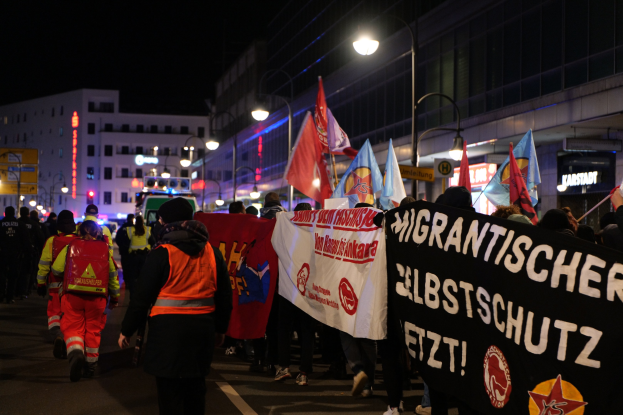 A group of people marching at night in Berlin, carrying flags and banners, with buildings, light poles, and a vehicle visible in the background.
