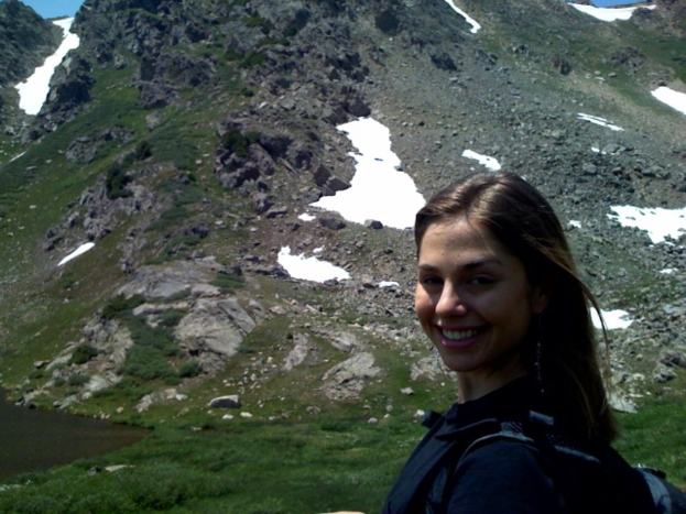 Eine Frau mit einer Tasche steht vor einem grasbewachsenen und teilweise schneebedeckten Felsen.