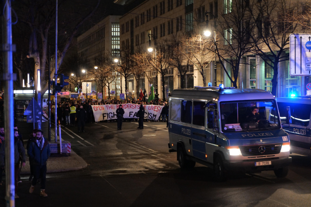 Gruppe von Menschen, die nachts eine von Straßenlaternen beleuchtete Straße entlanggehen, mit einem geparkten Polizeiwagen im Vordergrund, Bäumen und Gebäuden an der Straße und einem Banner im Hintergrund.