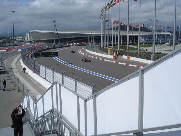 Eine Gruppe von Menschen auf einer Rennstrecke mit fahrenden Fahrzeugen drumherum, Kameras links, geflaggte Pfosten rechts und einem Stadion, Bäumen, Gebäuden und einem klaren blauen Himmel im Hintergrund.