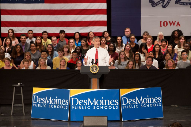 Ein Mann in einem weißen Hemd und roter Krawatte steht hinter einem Rednerpult mit drei Tafeln, auf denen Text steht, einem Hocker mit Mikrofon und einer sitzenden Menge im Hintergrund, mit einer Flagge und zusätzlichem Text.