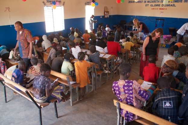Kinder auf Bänken in der Vordergrund mit Erwachsenen, die sie führen, Stühle, eine Tür, ein Fenster und ein Tisch im Hintergrund, Ballons und Text oben.