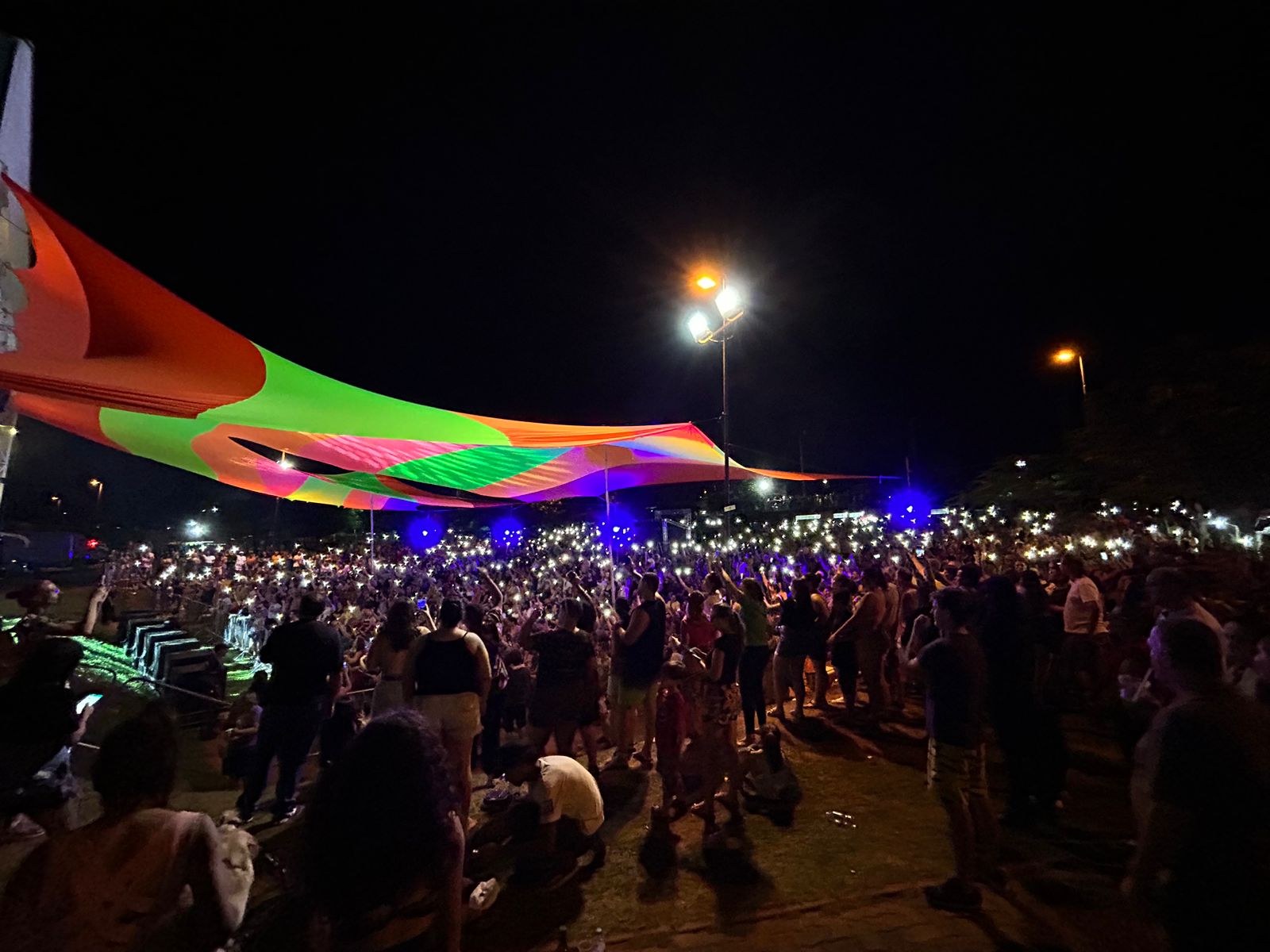 Eine große Menschenmenge steht vor einem Zelt in der Nacht, beleuchtet von bunten Lichtern und umgeben von beleuchteten Pfählen, mit einem Gebäude auf der linken Seite und dem Nachthimmel im Hintergrund während des Festivals der Künste im Jahr 2018.