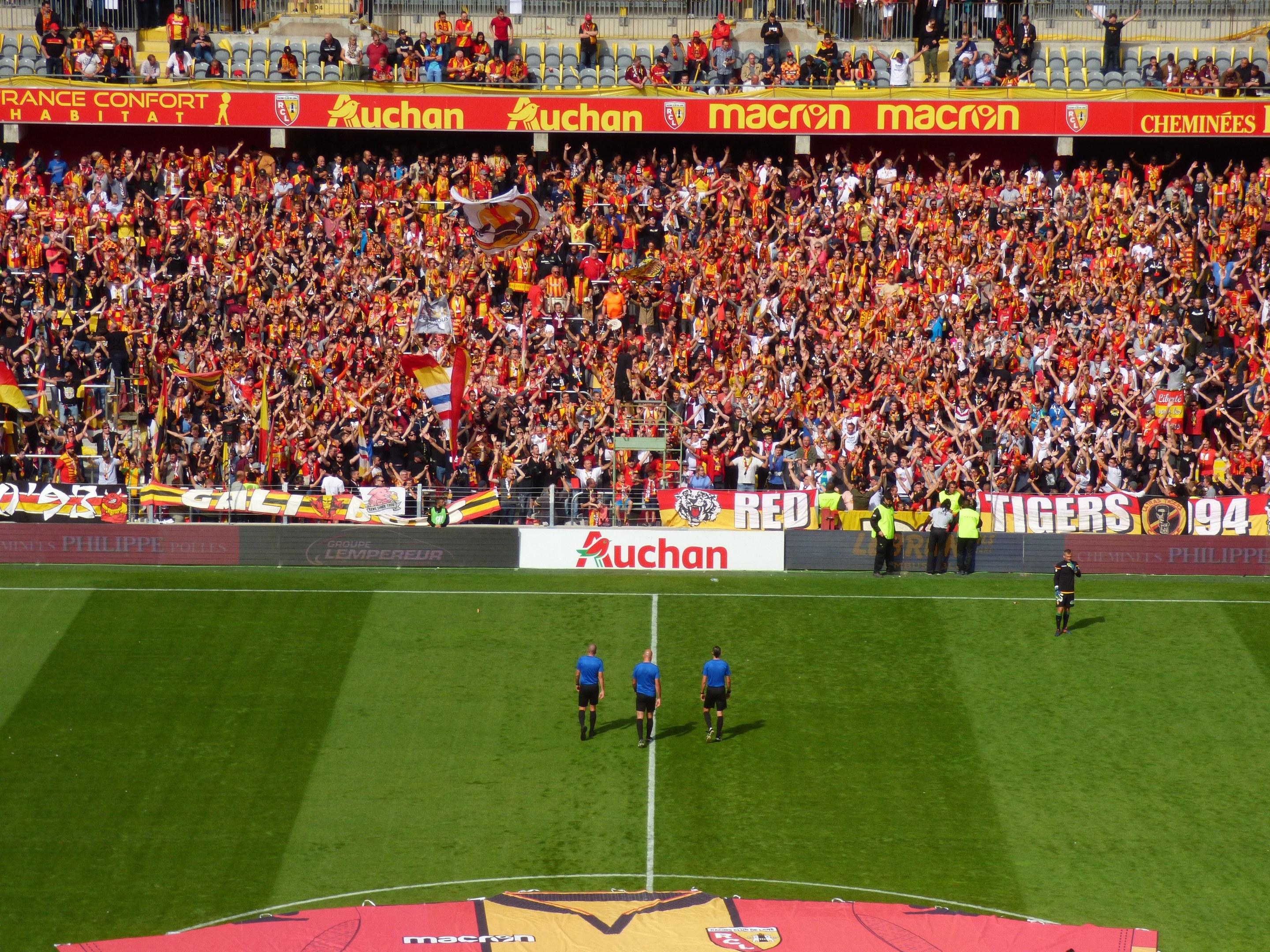 Ein Fußballspiel in einem Stadion mit Spielern auf dem Rasen und Zuschauern im Hintergrund, mit Schildern, Geländern und Treppen.