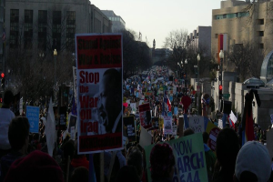 Eine Gruppe von Menschen demonstriert auf einer Straße, hält Plakate, mit Gebäuden, Bäumen und Laternen im Hintergrund unter einem klaren Himmel.