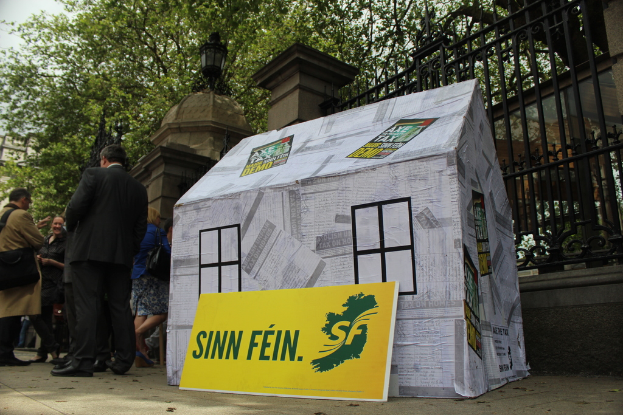 Ein Zelt und ein Werbeschild liegen auf dem Boden, umgeben von einer Menge, während im Hintergrund ein Eisenzaun, Säulen, Straßenlaternen, Bäume und der Himmel zu sehen sind.