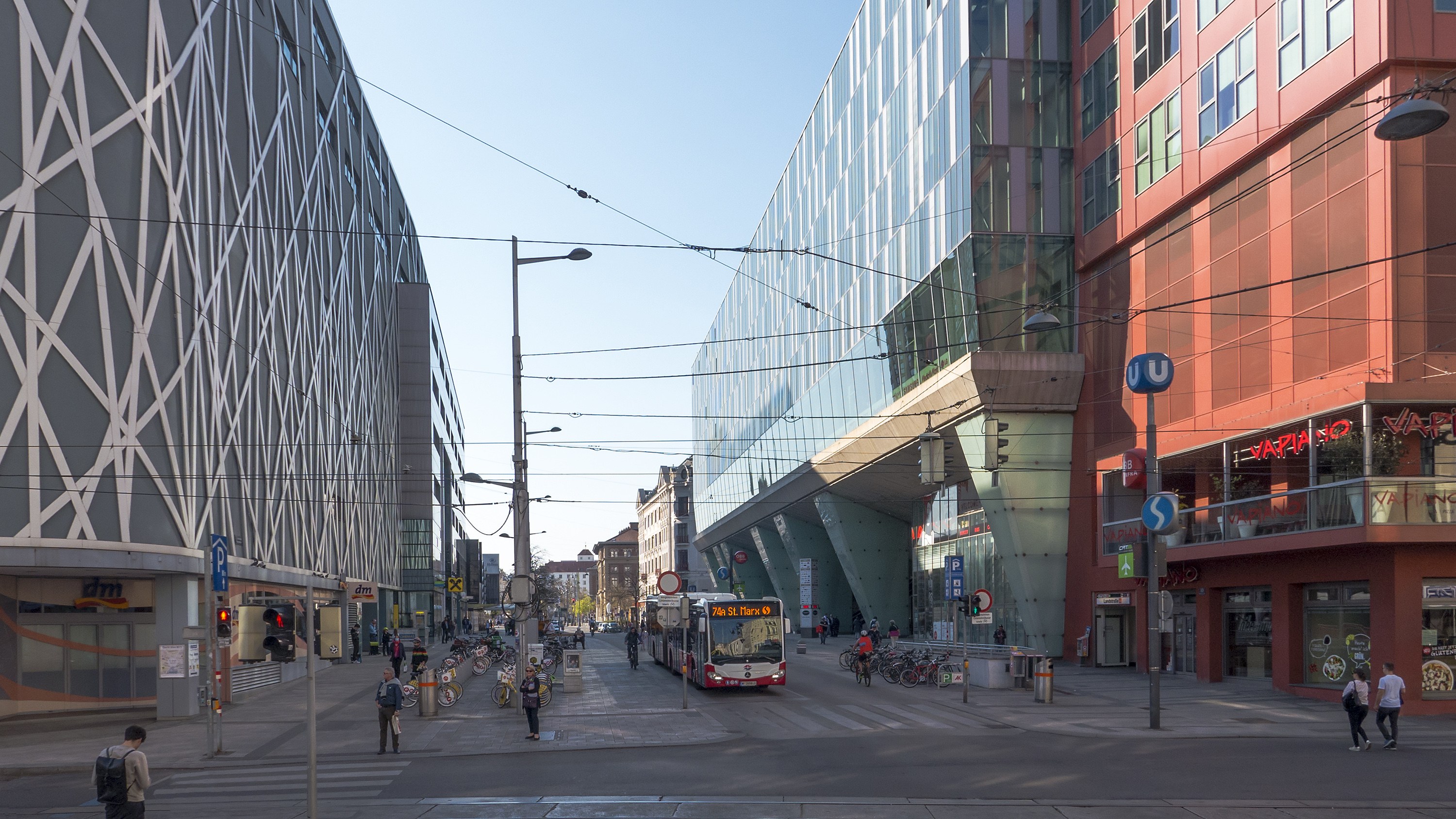 A busy city street with tall buildings, street infrastructure, vehicles, pedestrians, and a clear blue sky.