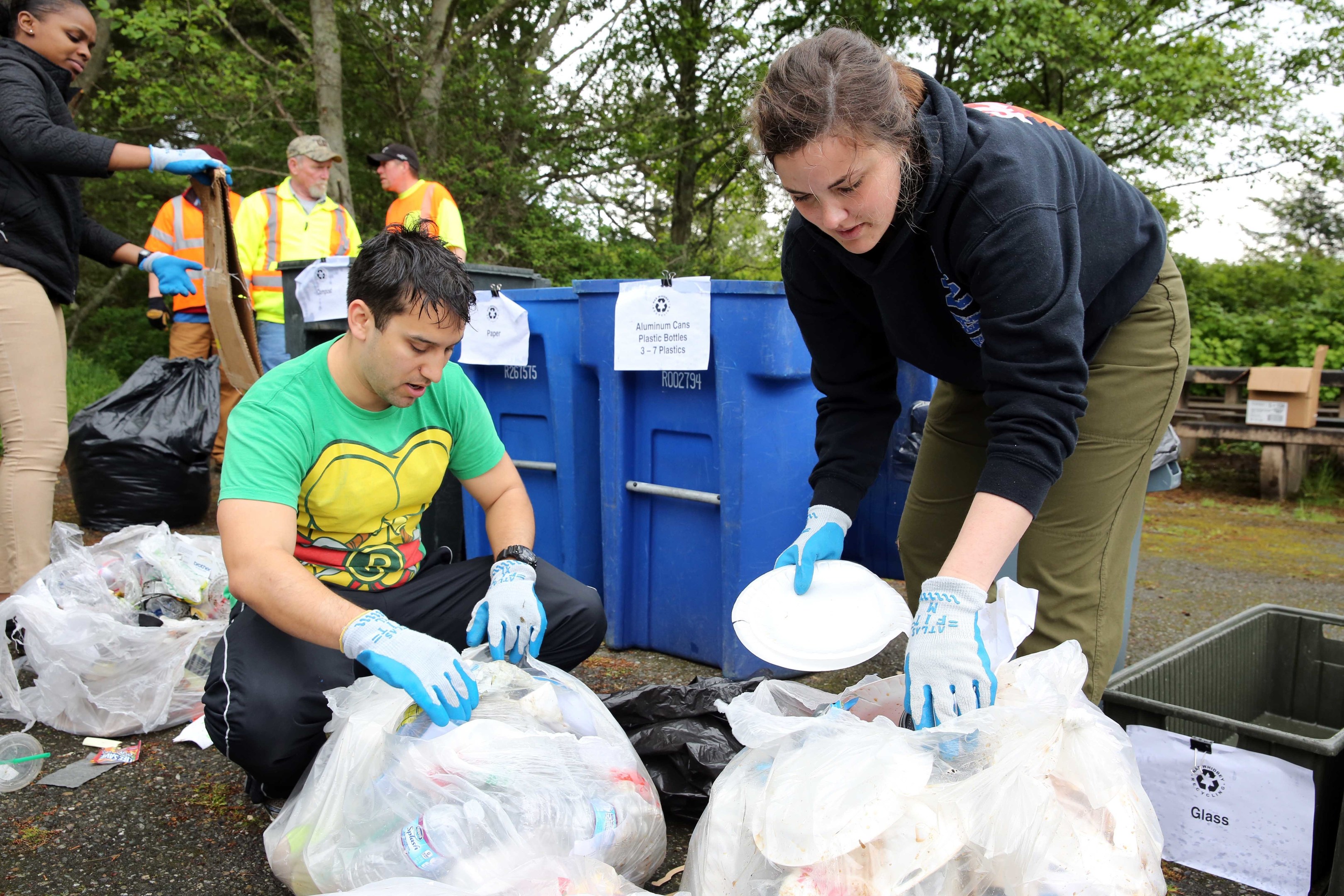 Two people in gloves collect litter on plates in a park, surrounded by trash and debris, with a dustbin and bench nearby under trees and a blue sky.