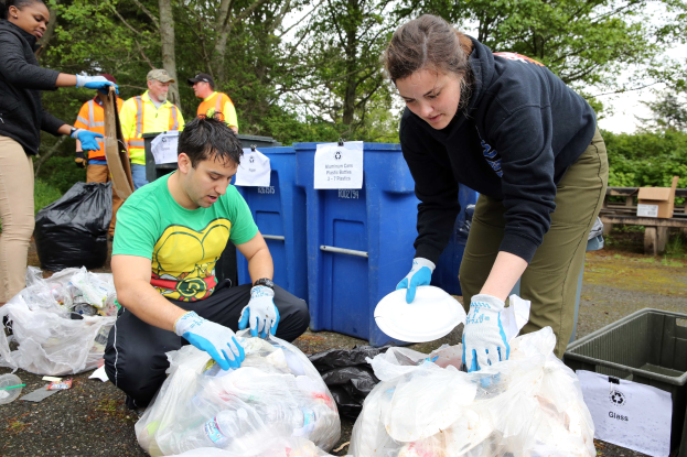 Two people in gloves collect litter on plates in a park, surrounded by trash and debris, with a dustbin and bench nearby under trees and a blue sky.