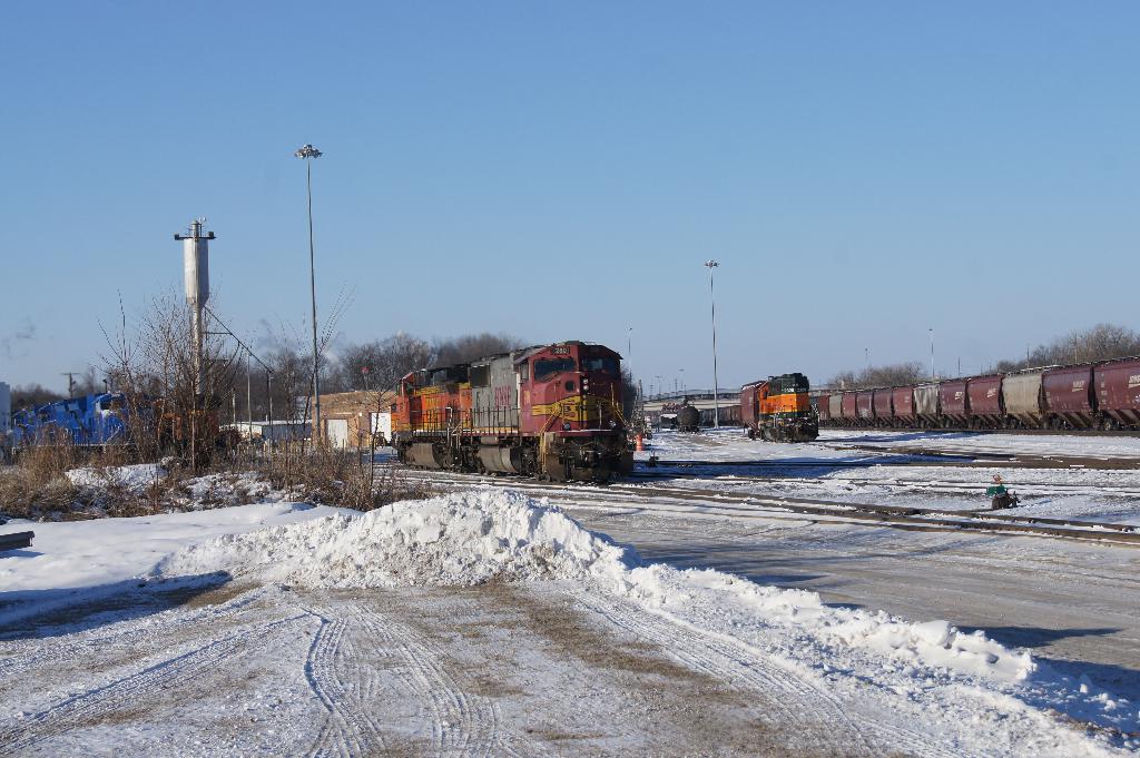 Eine Lokomotive vorne mit einem Waggon auf der rechten Seite, auf Schienen geparkt mit Schnee am Boden, trockenen Bäumen und einem Strommast im Hintergrund.