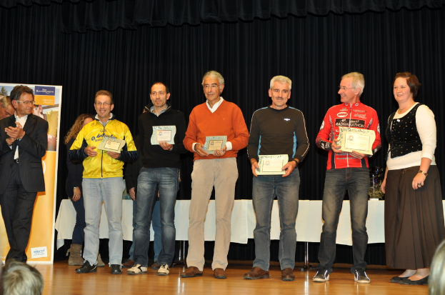 Eine Gruppe von Menschen steht auf einer Bühne und hält Zertifikate in den Händen, im Hintergrund eine Fahne, daneben ein Tisch und ein schwarzer Vorhang.