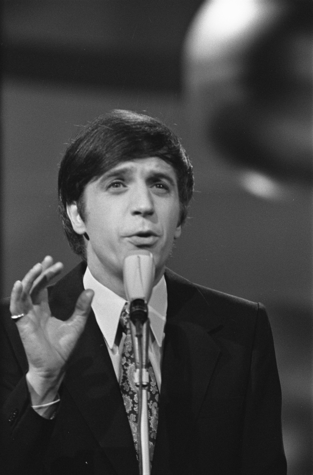 Ein Mann in einem schwarzen Anzug, weißem Hemd und gemustertem Schlips steht vor einem unscharfen Hintergrund und spricht in ein Mikrofon, mit ernster Miene und zurückgekämmtem Haar.