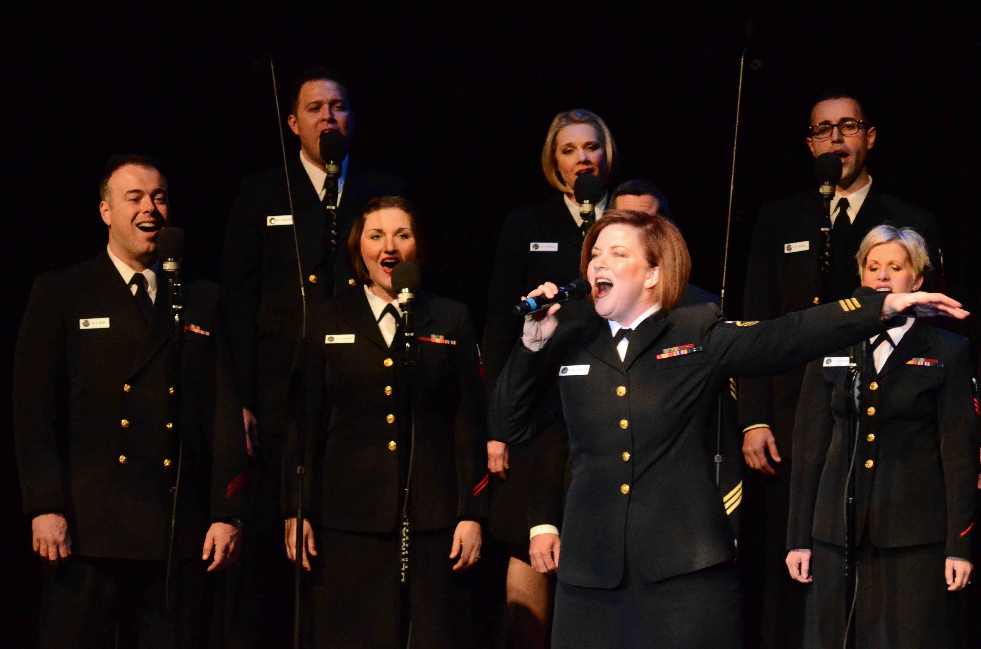 A group of people in military uniforms singing on a stage with microphones and stands against a dark background.