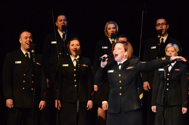 A group of people in military uniforms singing on a stage with microphones and stands against a dark background.