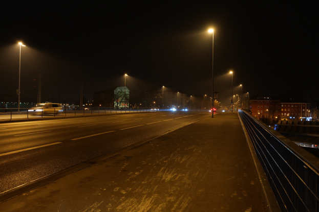Langzeitbelichtung einer nächtlichen Stadtstraße mit fahrenden Fahrzeugen, beleuchteten Laternen, Gebäuden, Bäumen, Geländern und einem dunklen Himmel.