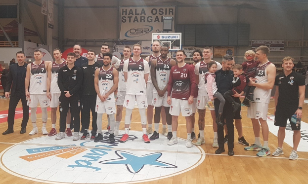 Mannergruppe mit Kindern auf einem Basketballfeld mit Schildern "Hala Osir Starga", Zuschauern und Basketballausrüstung im Hintergrund.