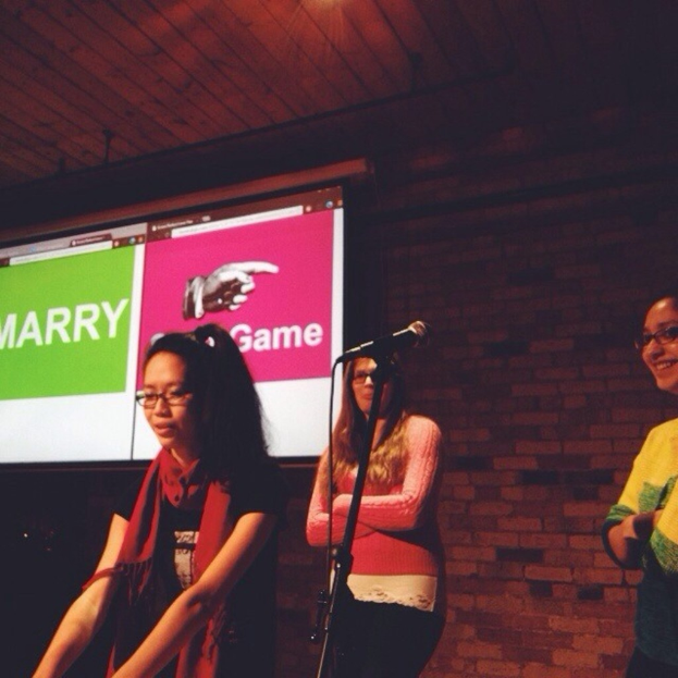 Drei Frauen stehen mit dem Gesicht nach links, mit einem Mikrofonständer in der Mitte und einem Bildschirm mit Text auf der linken Seite, vor einer Wand im Hintergrund.