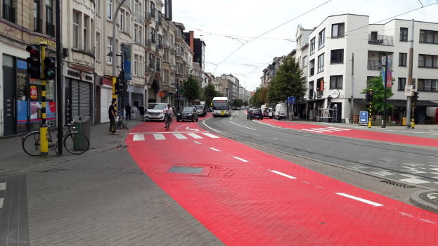 Rote Fahrradspur neben einer Stadtstraße mit fahrenden Fahrzeugen, Fußgängern auf dem Gehweg, Gebäuden, Bäumen und einem klaren blauen Himmel.