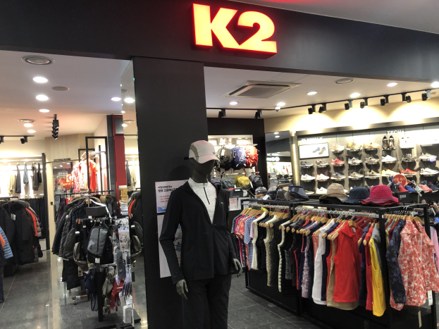 Interior of a K2 clothing store in Singapore displaying mannequins in caps, hanging clothes, shoe racks, and illuminated by overhead lights, with a promotional board on the wall.