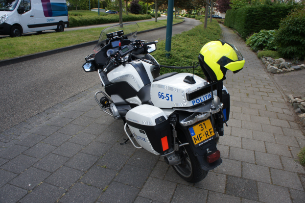 Eine Polizeimotorrad auf dem Seitenstreifen geparkt mit einem Helm darauf, umgeben von Bäumen, Polen, Gebäuden, Pflanzen, Gras, Steinen und anderen Fahrzeugen im Hintergrund.